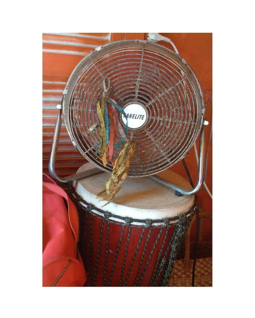 Chrome-finish fan sitting on a drum with hanging ribbons, against an orange wall, in an indoor setting with soft lighting.