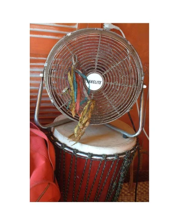 Chrome-finish fan sitting on a drum with hanging ribbons, against an orange wall, in an indoor setting with soft lighting.