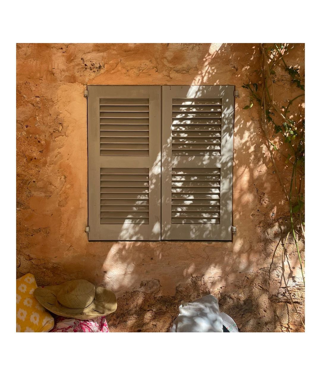 Exterior facade featuring a closed window with louvred shutters on a rustic wall, with shadows from foliage creating dappled light.