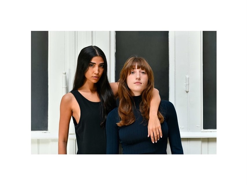 Two women pose indoors near a window with natural light. One woman has long dark hair, the other has long brown hair with bangs.