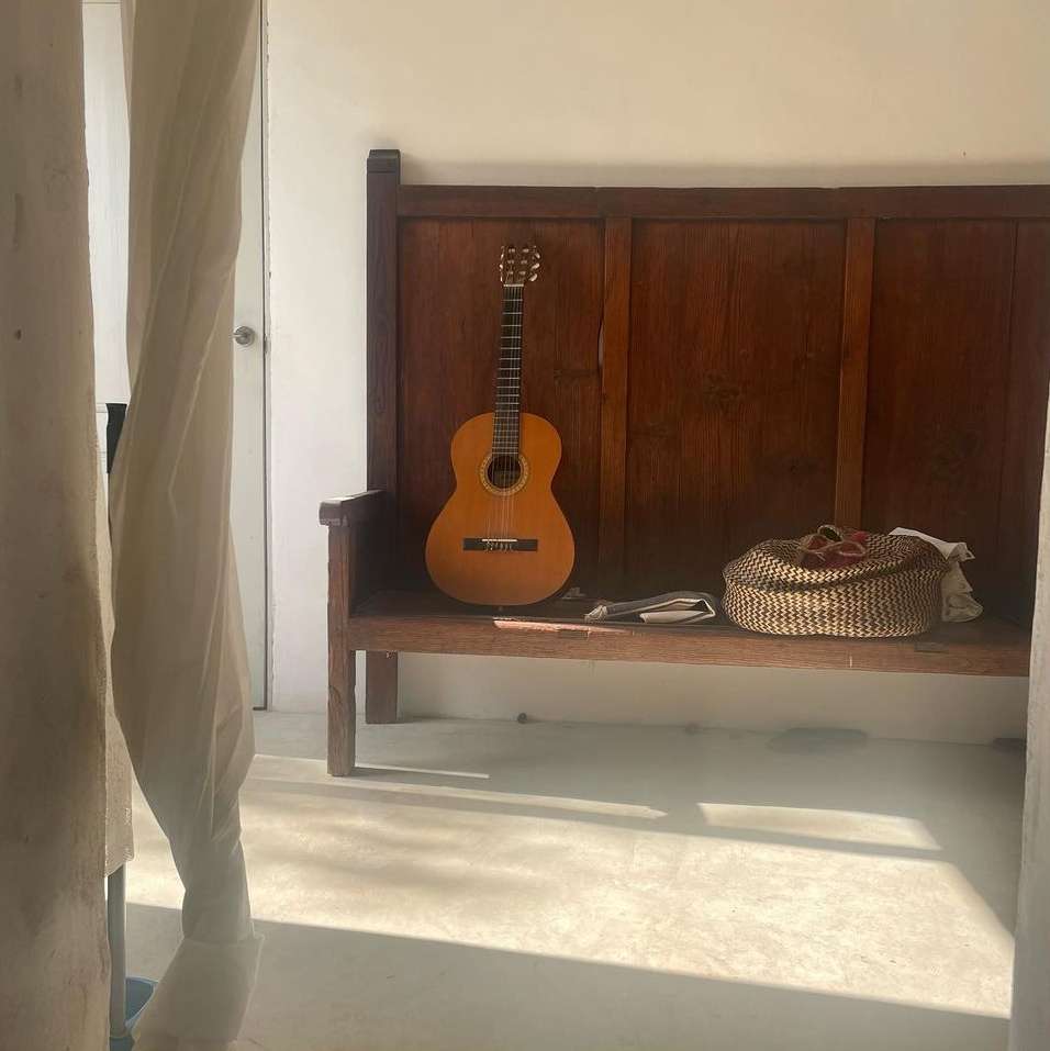 Acoustic guitar sits on a wooden bench indoors, accompanied by a woven basket and scattered papers in natural daylight, creating a serene scene.