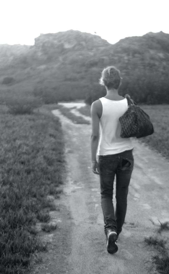 A woman walks away on a dirt path with a bag over her shoulder, heading towards the distant hills in a monochrome outdoor scene.