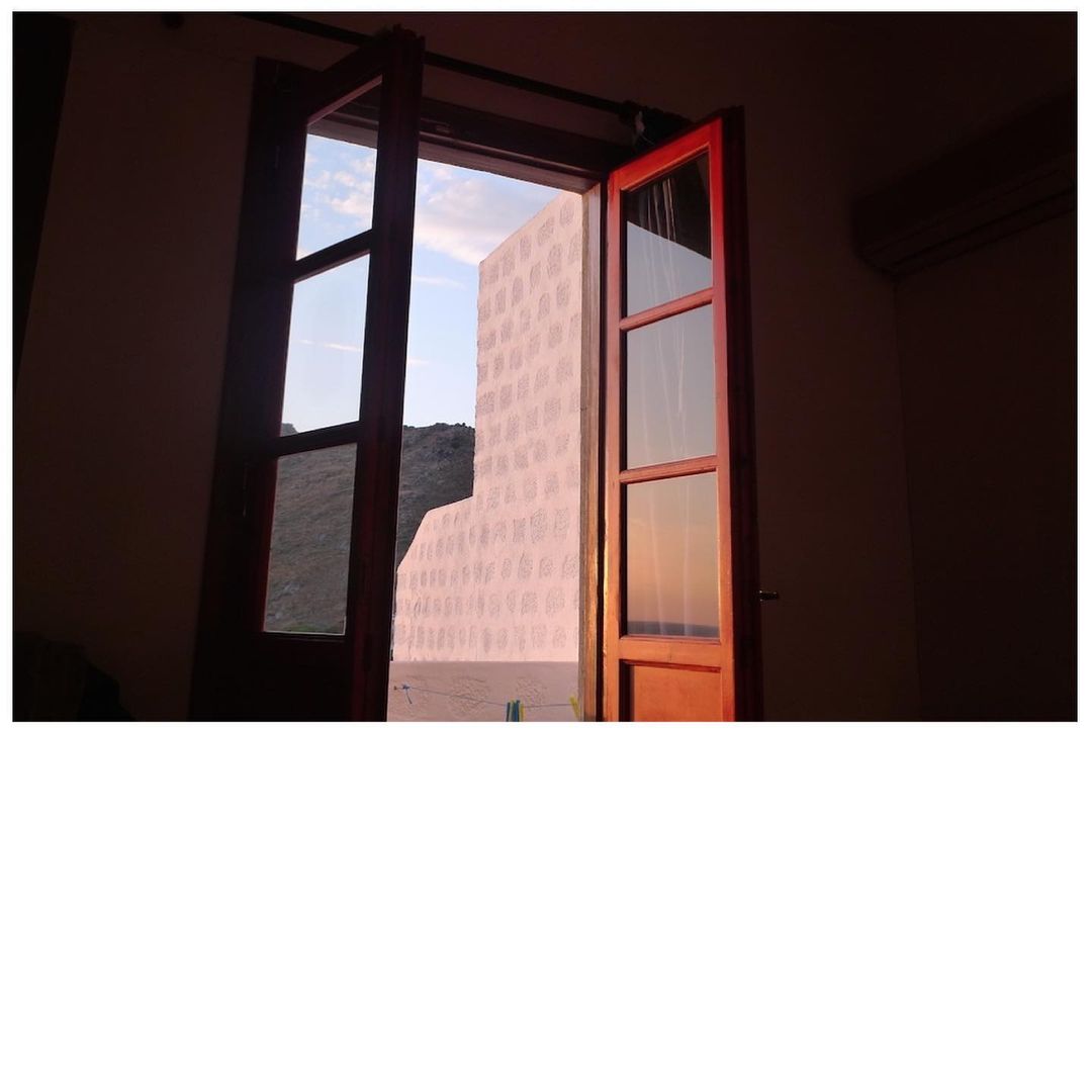 Open wooden window frame in a dimly lit room overlooking a bright outdoor scene with a patterned wall and distant hill.