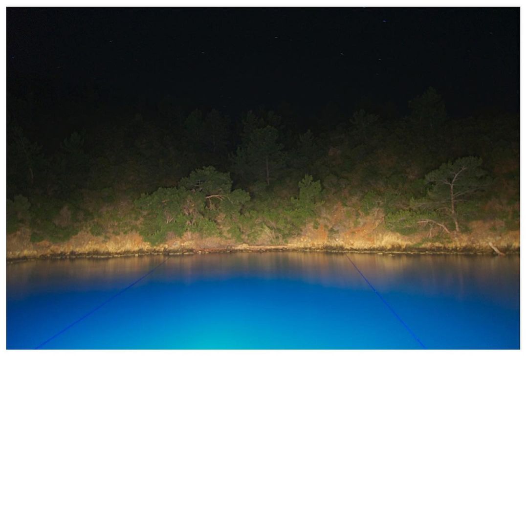 Nighttime view of a vibrant blue lake reflecting the sky and bordering trees along the shoreline under a dimly lit, starlit sky.