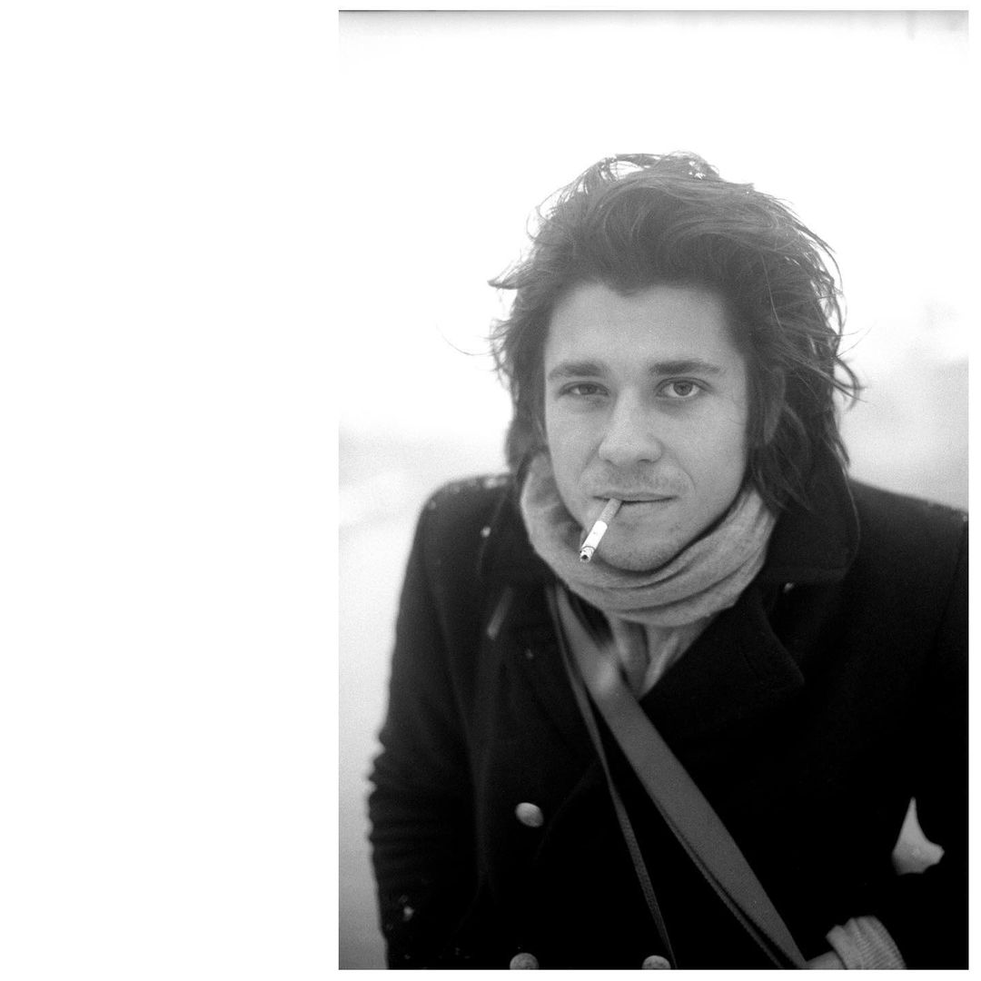 Black and white portrait of a man with long hair, wearing a coat and scarf, holding a cigarette to his lips while outside.