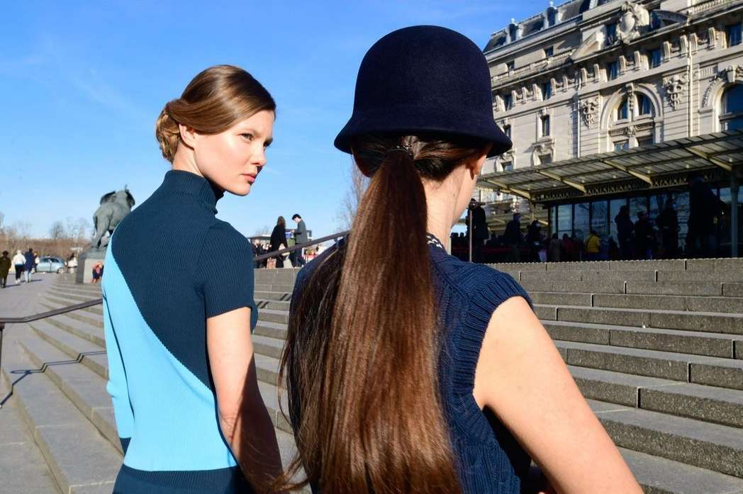 Two women on stone steps in sunlight; one in blue sweater, one in hat with long hair, building in background. Outdoor fashion shot.