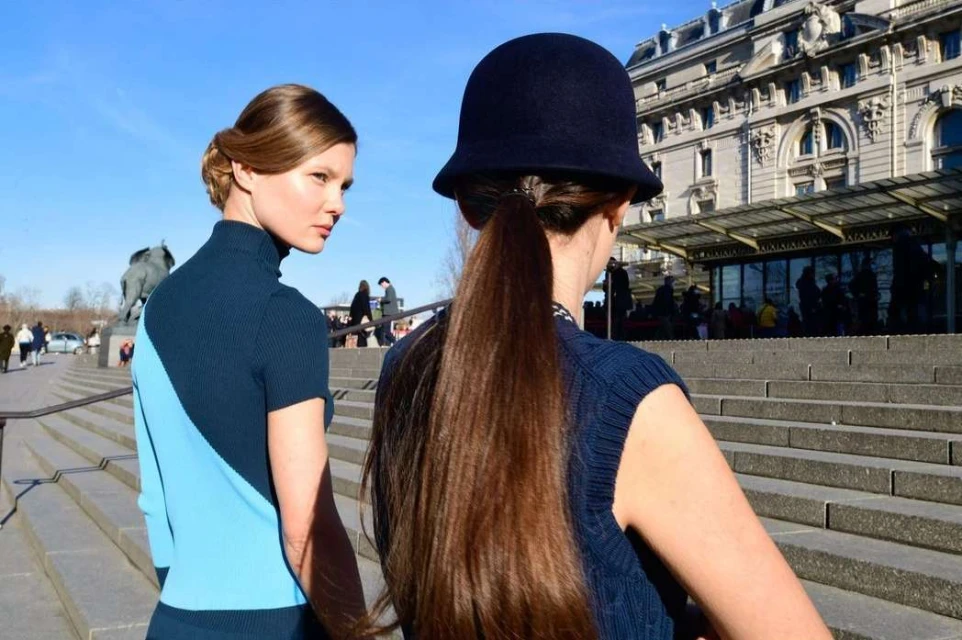 Two women on stone steps in sunlight; one in blue sweater, one in hat with long hair, building in background. Outdoor fashion shot.