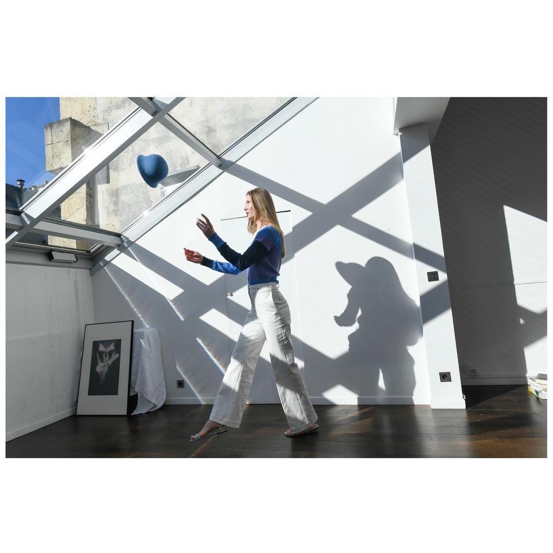 Woman tossing a blue ball indoors with natural light, wearing a two-tone blue top and white pants. Interior features white walls and wood floor.