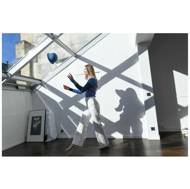 Woman tossing a blue ball indoors with natural light, wearing a two-tone blue top and white pants. Interior features white walls and wood floor.