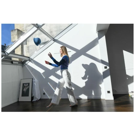Woman tossing a blue ball indoors with natural light, wearing a two-tone blue top and white pants. Interior features white walls and wood floor.