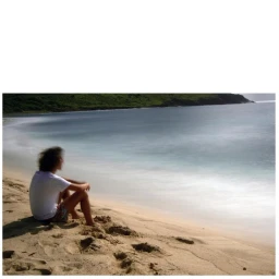 A person with dark hair seated on a sandy beach looking out toward the ocean under soft natural light.