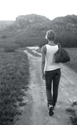 Person wearing a white tank top and dark jeans walking away along a rural dirt path toward hills