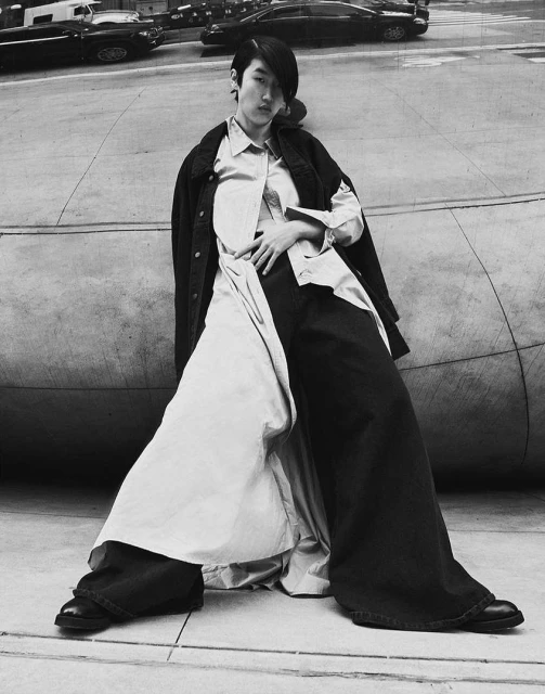 Monochrome portrait of a model in wide-leg trousers and a jacket posing outdoors with cars in the background, cool tones, editorial style.