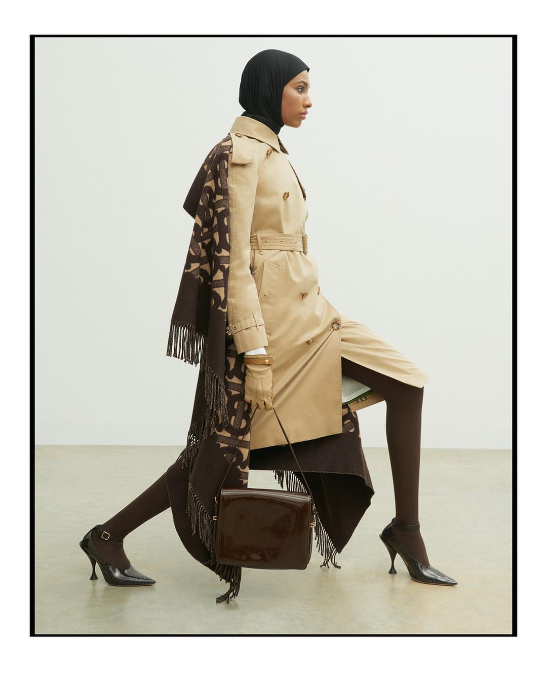 A person in a hijab and beige trench coat strikes a dynamic pose against a white backdrop with a brown patterned scarf and purse.