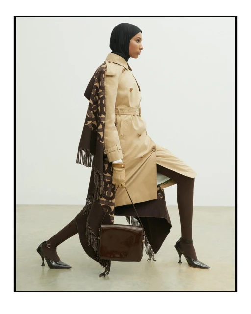 A person in a hijab and beige trench coat strikes a dynamic pose against a white backdrop with a brown patterned scarf and purse.