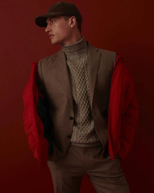 Man in a brown suit, cable-knit turtleneck sweater, and cap poses against a deep red background, with a red jacket draped over one arm.