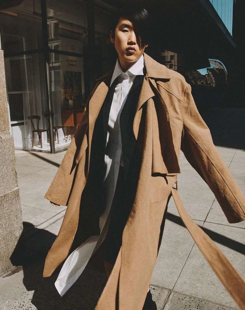 Adult fashion model wearing a tan trench coat and white shirt in an urban setting. Strong sunlight and shadows are present.