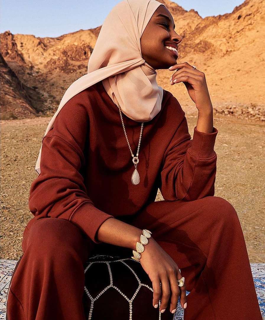 A smiling woman in a hijab and rust-colored sweatsuit sits in a desert landscape in a medium shot, accessorized with jewelry.