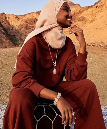 A smiling woman in a hijab and rust-colored sweatsuit sits in a desert landscape in a medium shot, accessorized with jewelry.
