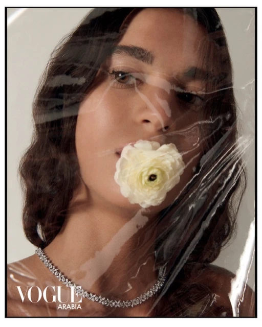 Close-up fashion portrait of a woman with flower near her mouth, wearing a diamond necklace and plastic wrap. Vogue Arabia logo is visible.