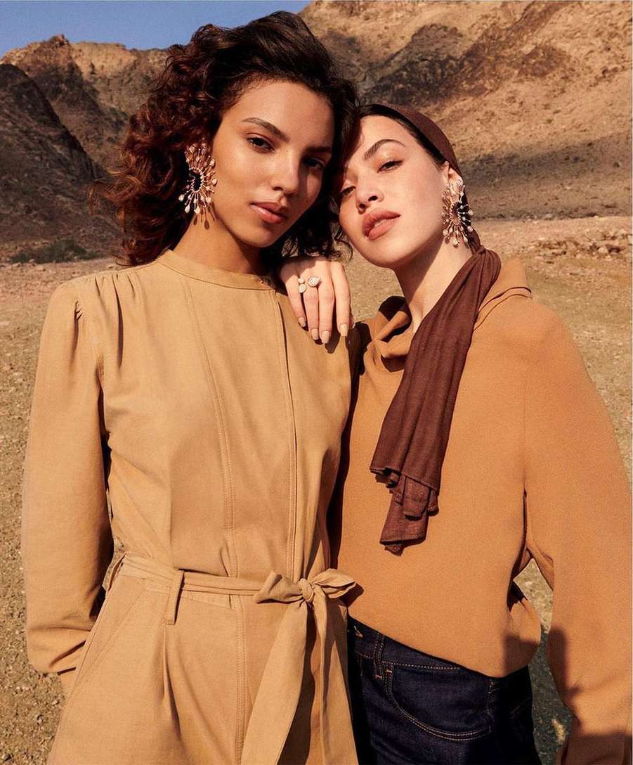 Two young adult women pose in earth-tone fashion in front of arid, mountainous landscape with clear sky for an outdoor fashion editorial.