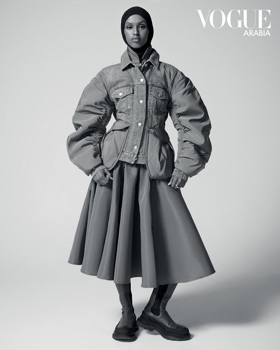 Full-length black and white portrait of a woman wearing a black headscarf, denim jacket, full skirt, and boots, posing in a photo studio.