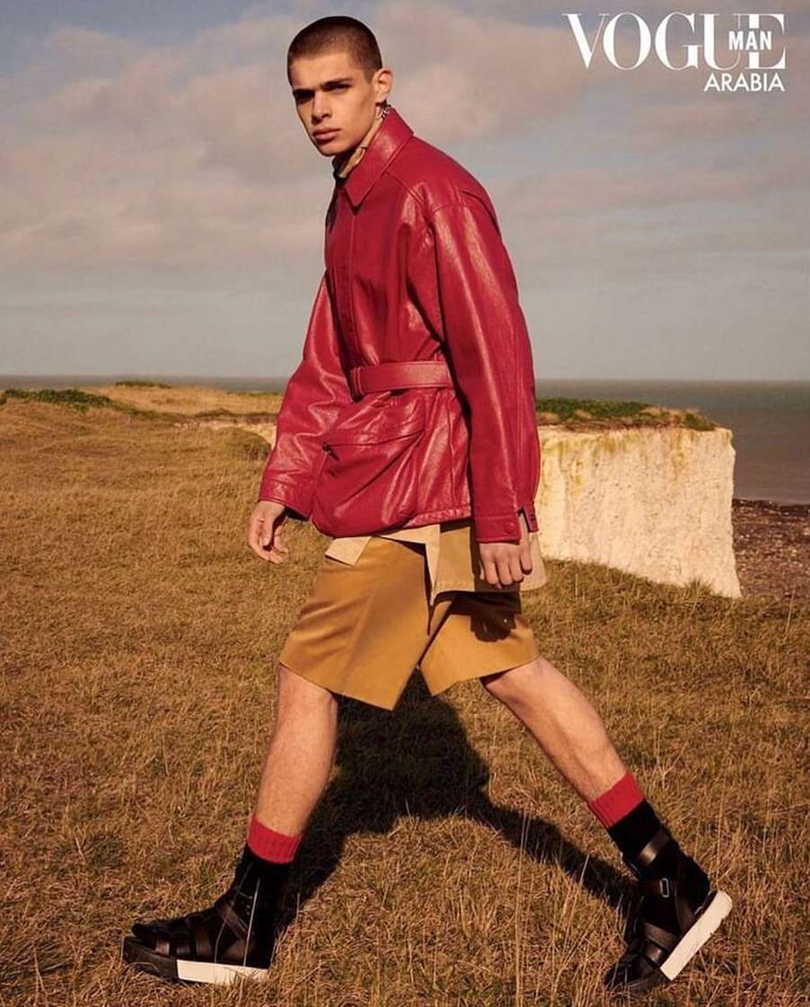 A fashionable man in a red jacket and tan shorts walks outdoors on a grassy field near a bluff, showcasing editorial style and Vogue Man Arabia.