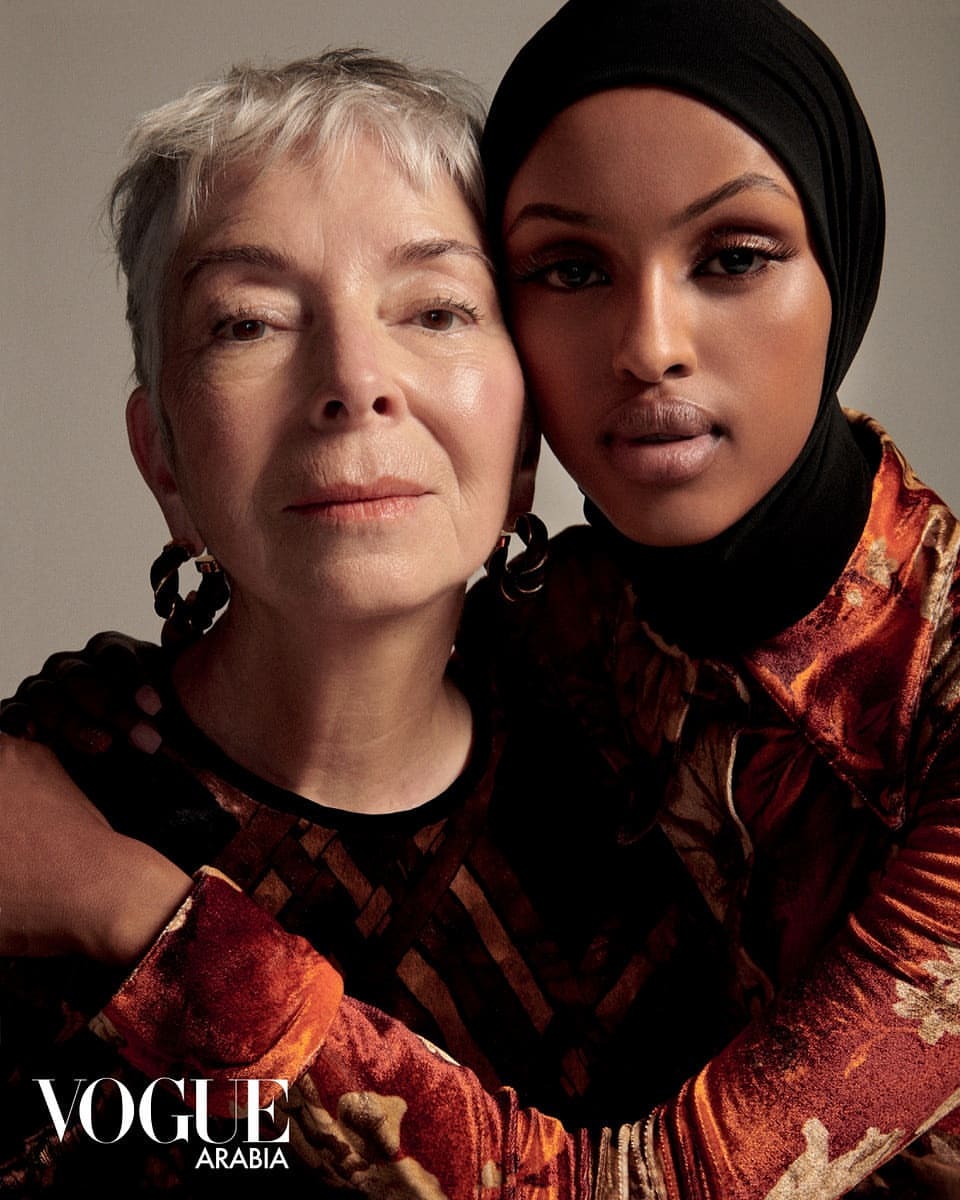 Close-up of two women with an arm around each other. One woman wears a hijab, the other has short grey hair. Vogue Arabia logo visible.