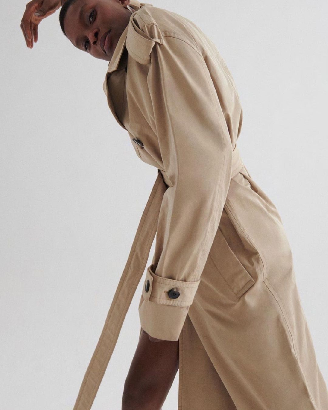 A tan-skinned person poses in a tan trench coat against a plain white backdrop, capturing a high-fashion aesthetic and minimalist style.