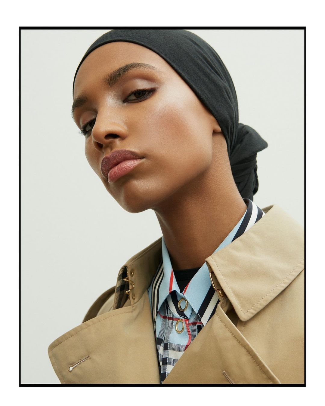 Portrait of a woman in a black headscarf and tan trench coat looking at the camera against a light neutral background.