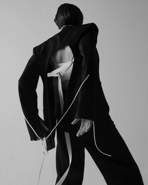 Back view of a fashion model wearing a black suit with white rope accents, with an open back, in a studio setting with soft lighting.