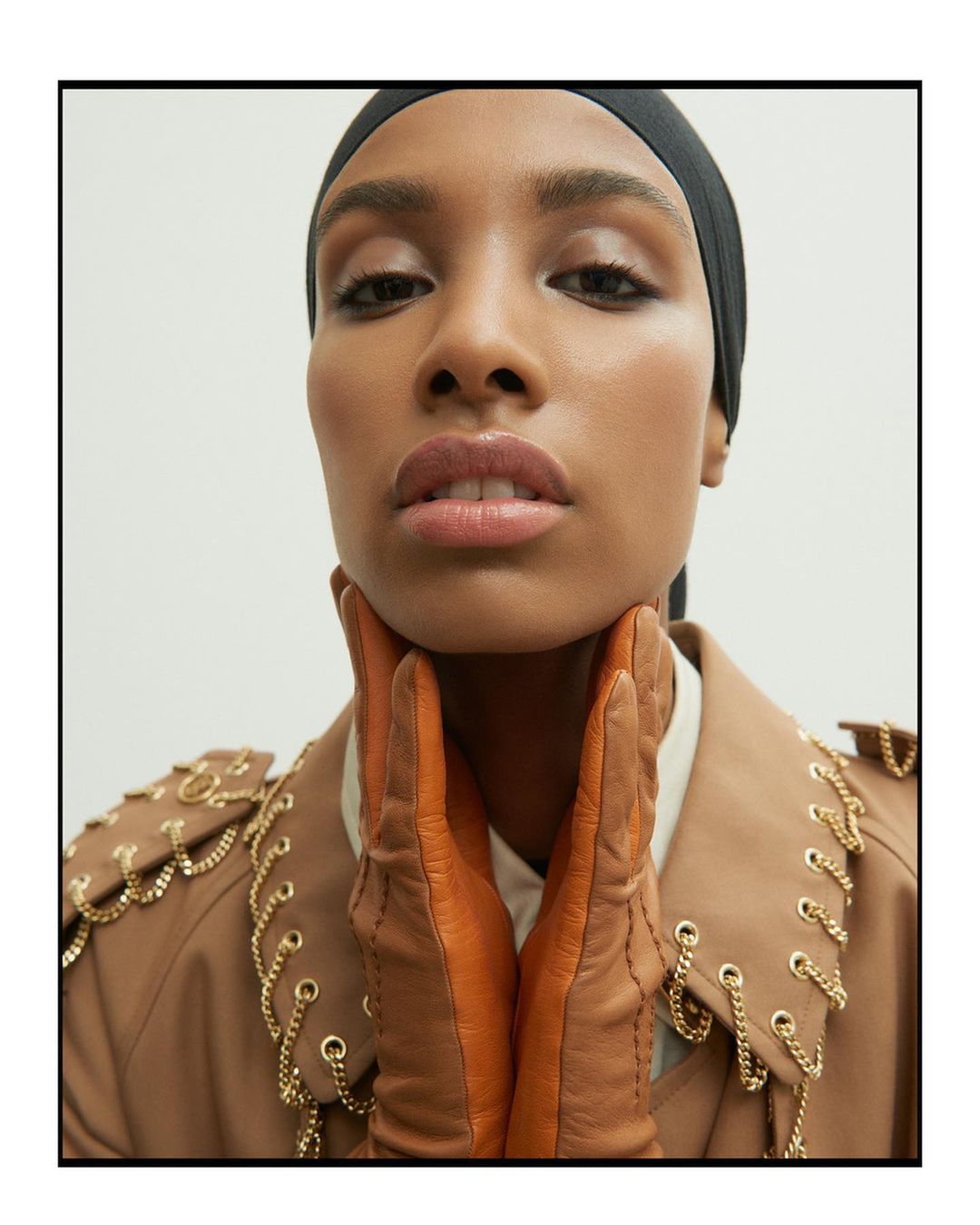Close-up of a woman with medium brown skin, wearing a black headscarf, tan coat with gold chains, and brown leather gloves.