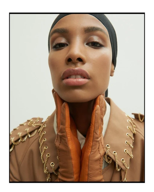 Close-up of a woman with medium brown skin, wearing a black headscarf, tan coat with gold chains, and brown leather gloves.