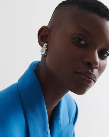 Close-up portrait of a Black woman with short hair wearing a blue blazer and an abstract silver earring, against a white background.