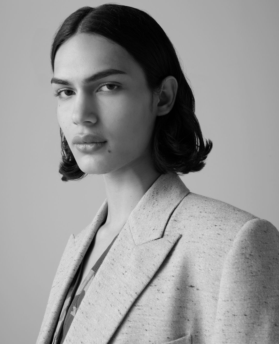 Black and white portrait of a person with short dark hair, wearing a speckled blazer and a patterned shirt, against a neutral backdrop.