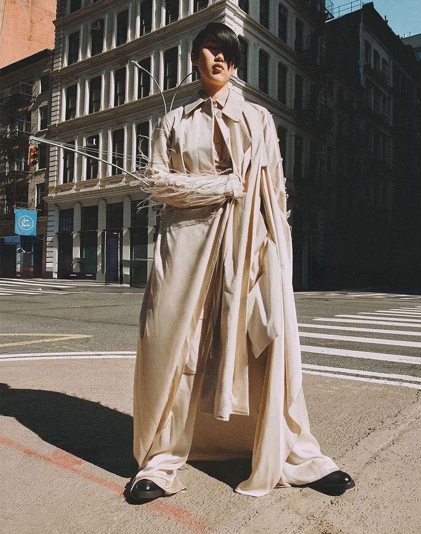 Woman wearing a long, light-colored trench coat and wide pants standing on a city street, buildings in the background.