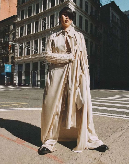 Woman wearing a long, light-colored trench coat and wide pants standing on a city street, buildings in the background.