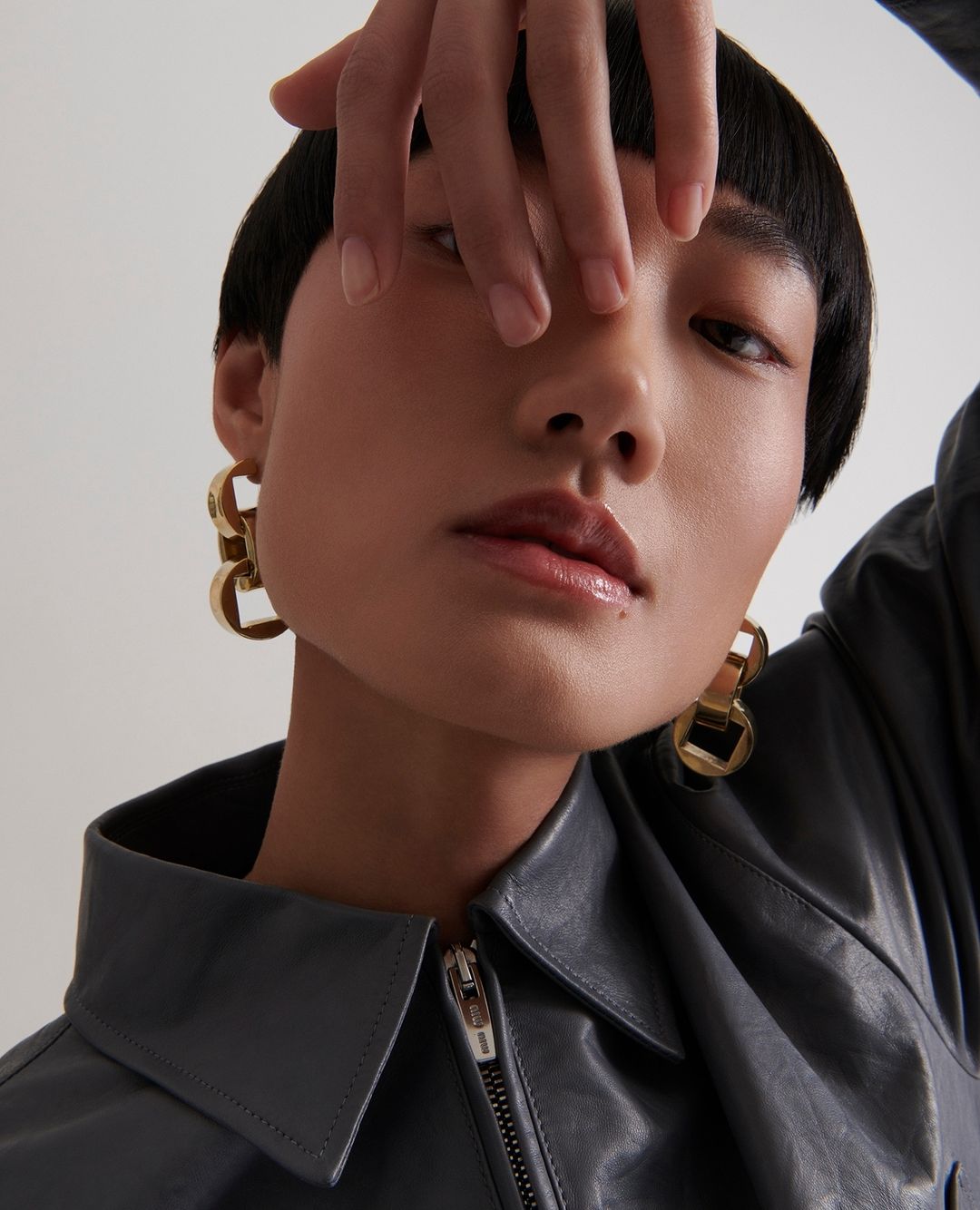 Close-up portrait of a young Asian woman with short black hair wearing a grey jacket and gold earrings against a light background.