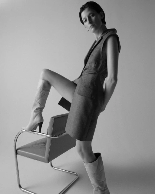 A woman strikes a fashionable pose, leaning against a chair with a sleeveless dress and high boots against a neutral backdrop.