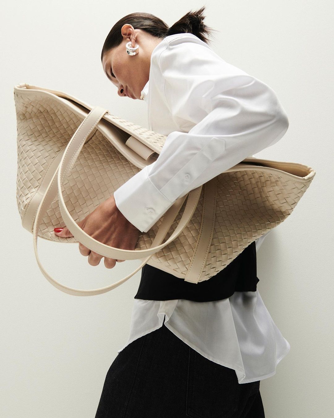 Woman holds a woven beige tote bag. She wears a white button-down shirt and black skirt against a neutral background in a studio shot.