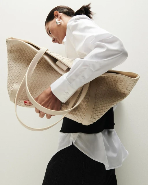 Woman holds a woven beige tote bag. She wears a white button-down shirt and black skirt against a neutral background in a studio shot.