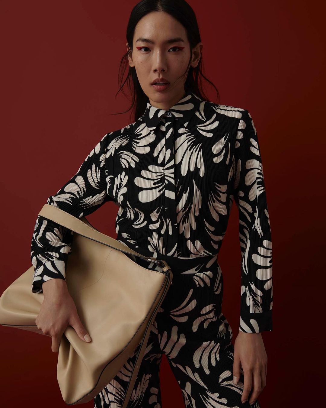An Asian woman in a patterned shirt and pants holds a beige bag against a red background, showcasing fashion and style.