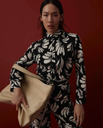 An Asian woman in a patterned shirt and pants holds a beige bag against a red background, showcasing fashion and style.