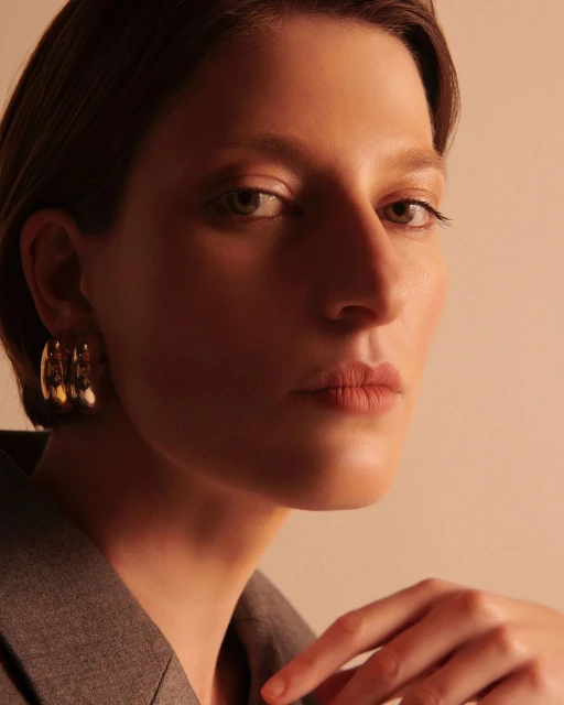 Close-up portrait of an adult female with gold hoop earrings wearing a grey jacket in a warm, softly lit indoor environment.