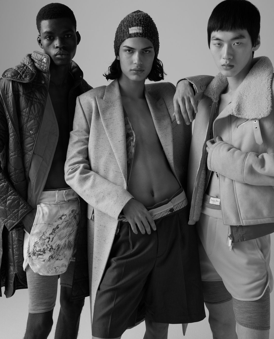 Three men are posing indoors against a plain background. They are wearing jackets, shorts, and hats. Black and white fashion photo.