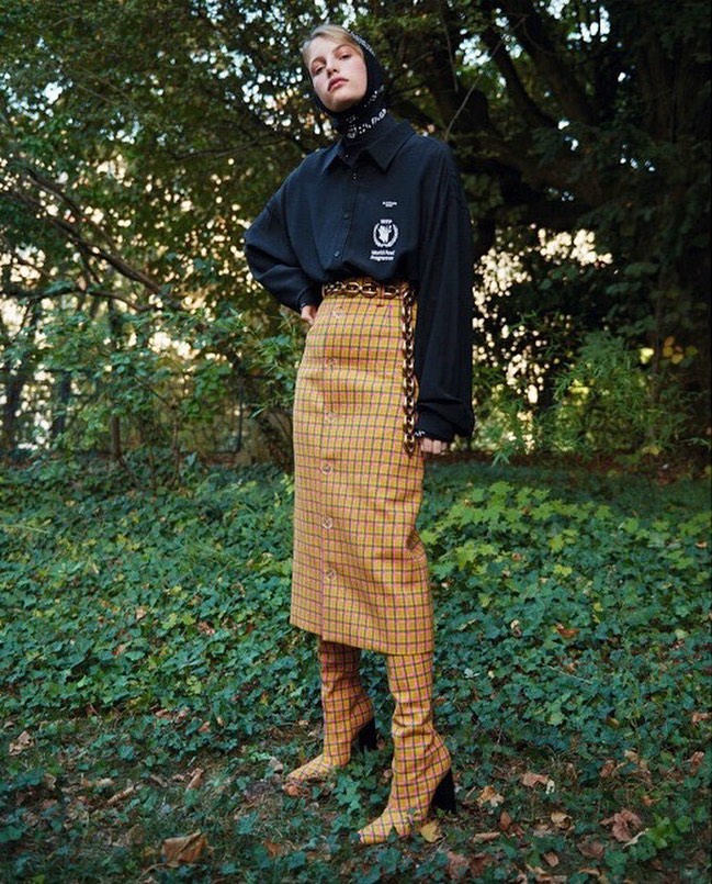 Full-length shot of a fashionable woman in a button-down shirt, plaid skirt, and heeled boots standing in a grassy outdoor area with trees.