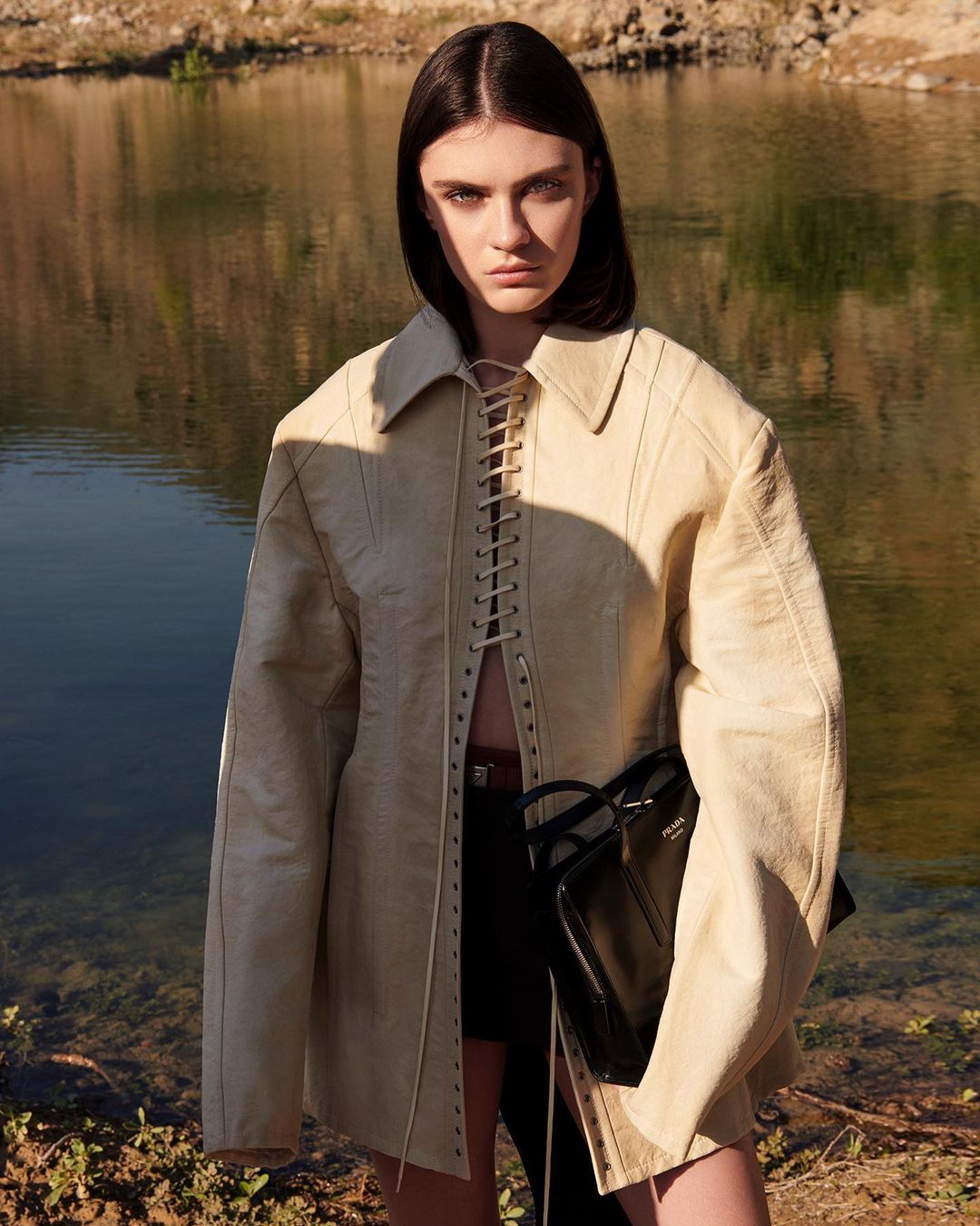 Model with dark bob hairstyle wearing a cream lace-up jacket holding a black handbag outdoors by a lake.