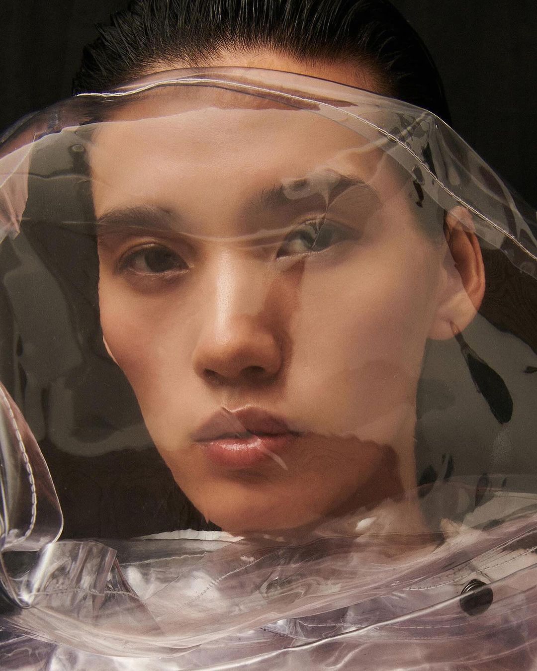 Headshot of a young adult woman with dark hair under a clear plastic sheet, facing the camera in a dark studio setting.