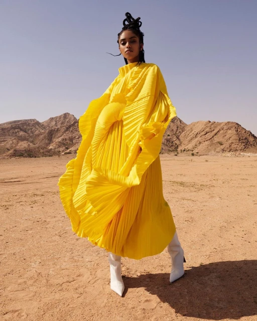 Fashion editorial of a young adult woman in a yellow dress and white boots posing in a desert environment with mountains in the background.