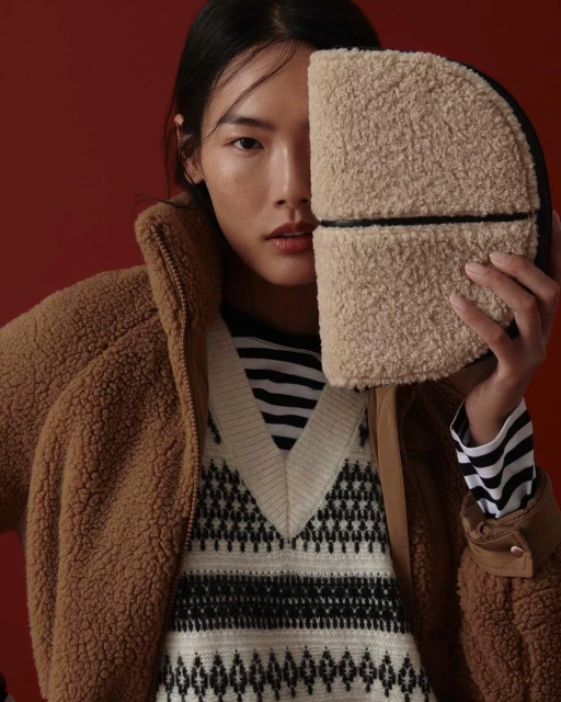Fashion portrait of an Asian woman in a patterned vest and jacket, holding a sherpa handbag against a dark red background.